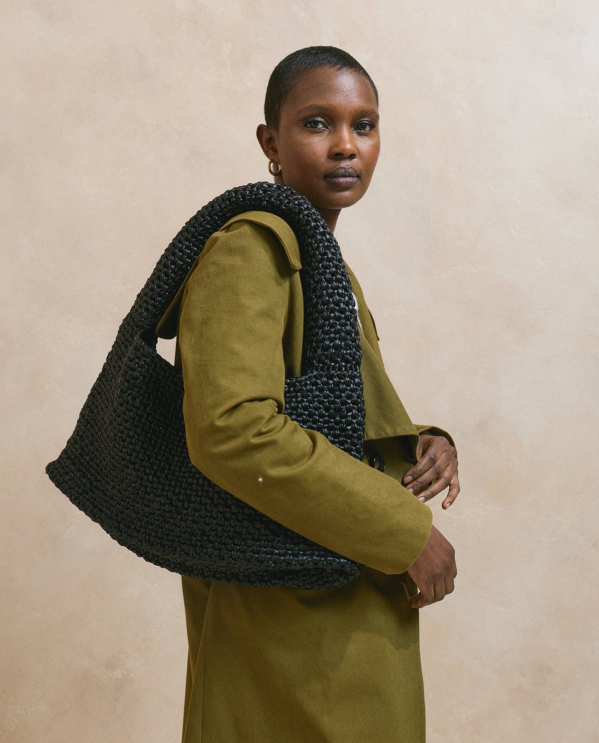 Mala preta trançada com alça larga sobre o ombro de uma mulher com casaco verde.