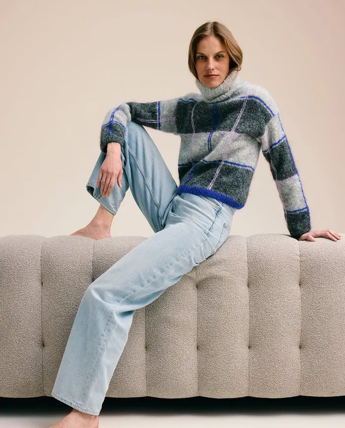 Mulher vestindo camisola xadrez cinza, azul e preta e calças de ganga azuis, sentada num sofá cinza claro.