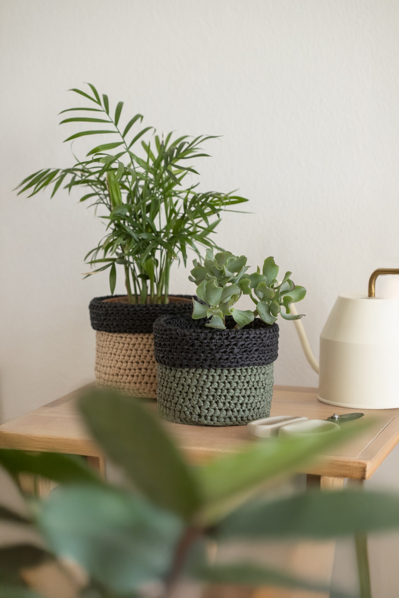 Duas plantas em vasos bicolores de crochê sobre mesa de madeira ao lado de regador e tesoura
