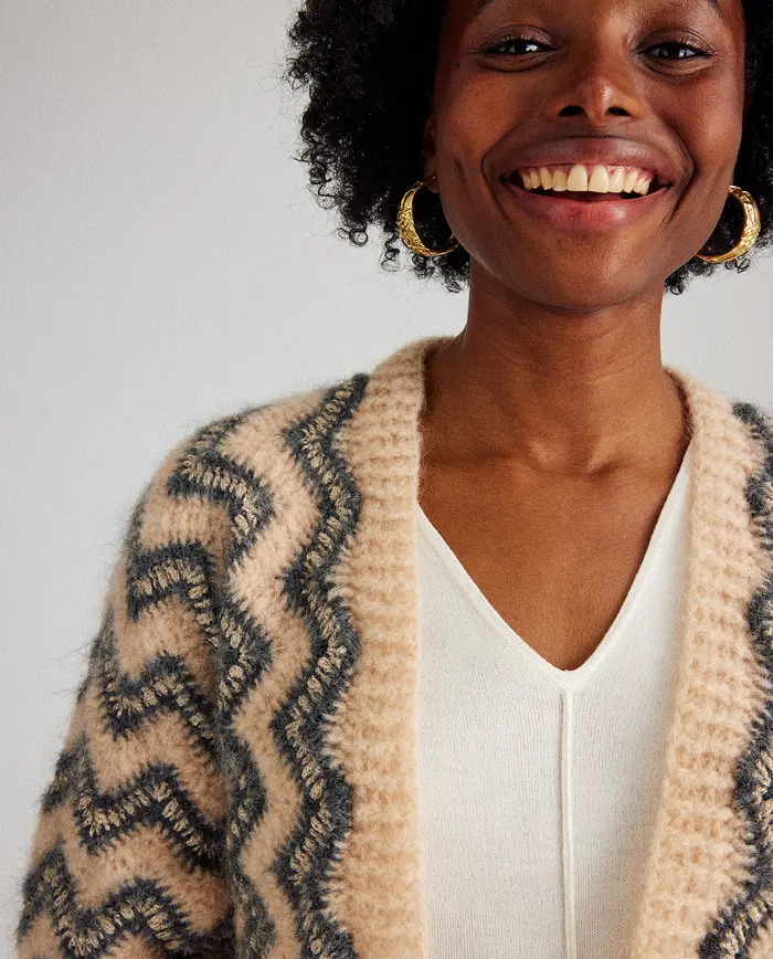 Mulher com casaco bege de malha felpuda com padrão zigzag cinza e blusa branca