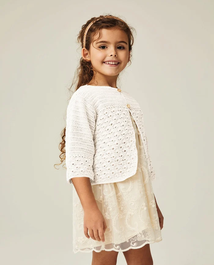 Menina com casaco branco de malha, vestido creme bordado e diadema rosa