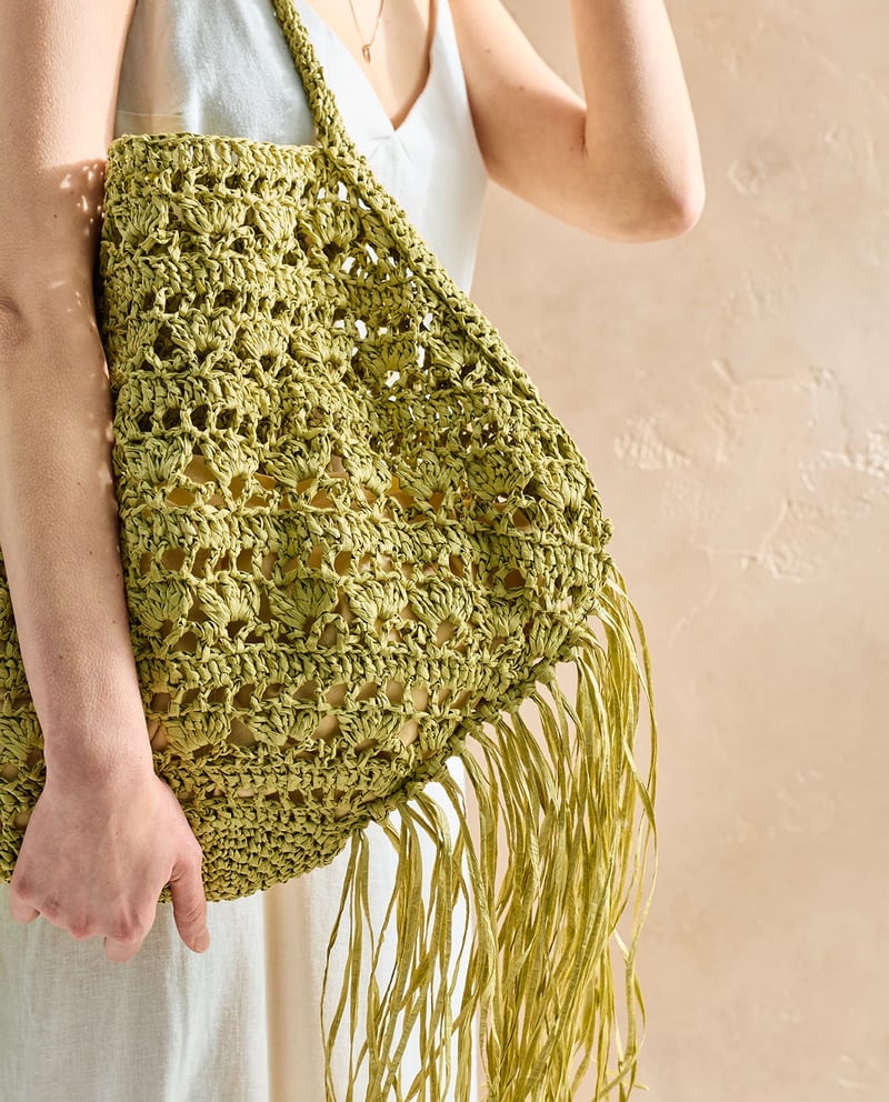 Mala de ombro verde claro em crochet com franjas, usada por mulher com vestido branco.