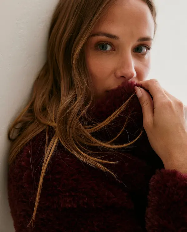 Casaco felpudo vinho usado por mulher junto à parede branca