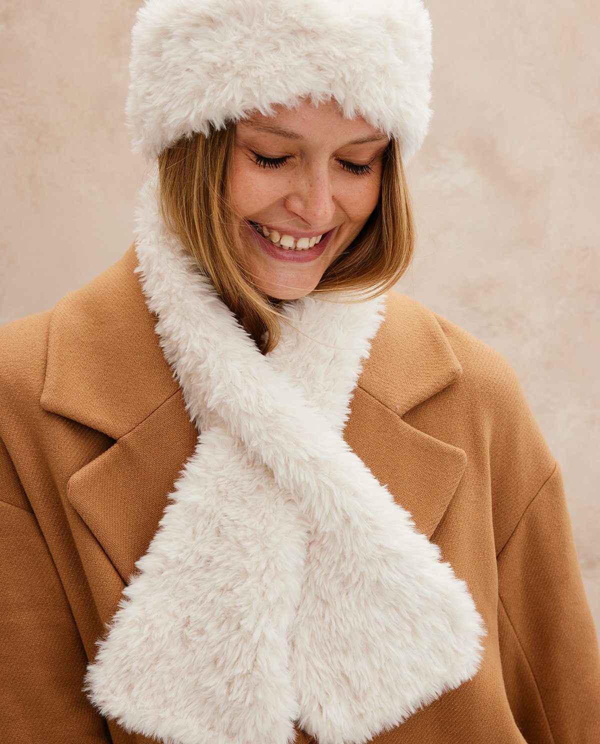 Mulher sorridente com chapéu e cachecol branco de pelo sintético e casaco camel.