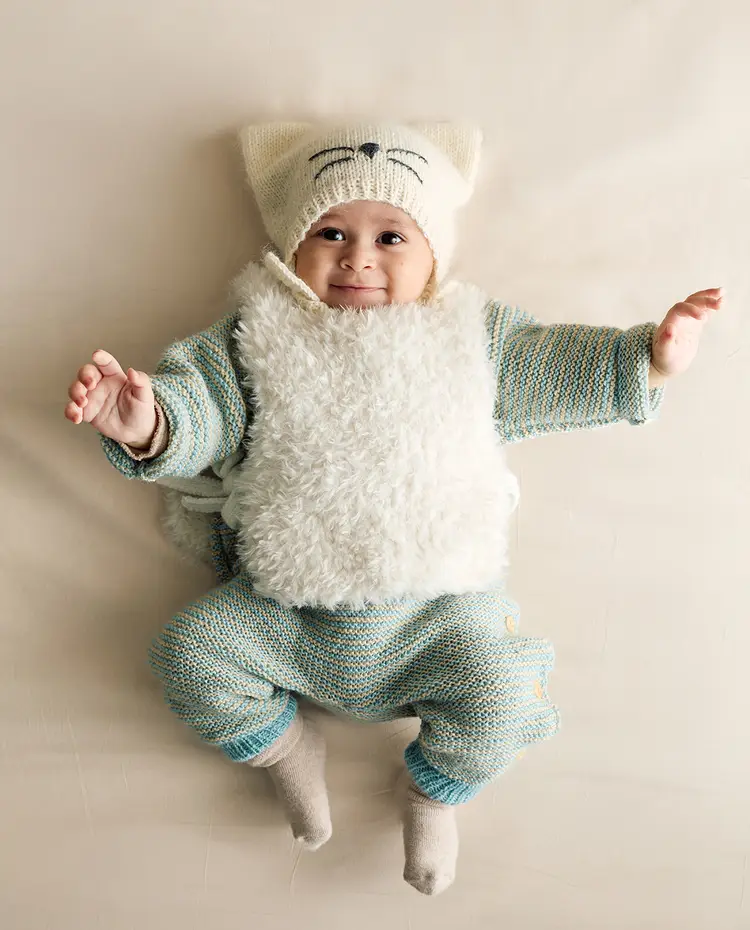 Bebé deitado com roupa de malha azul com creme, colete branco felpudo e gorro creme com rosto de gato