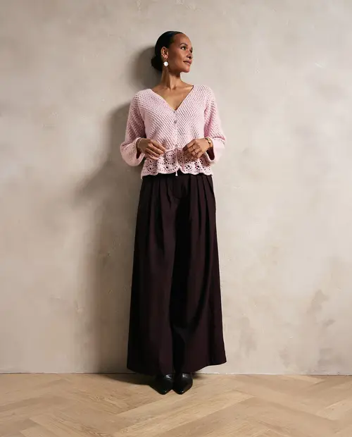 Mulher com camisola rosa de malha e calças largas pretas junto a uma parede cinzenta