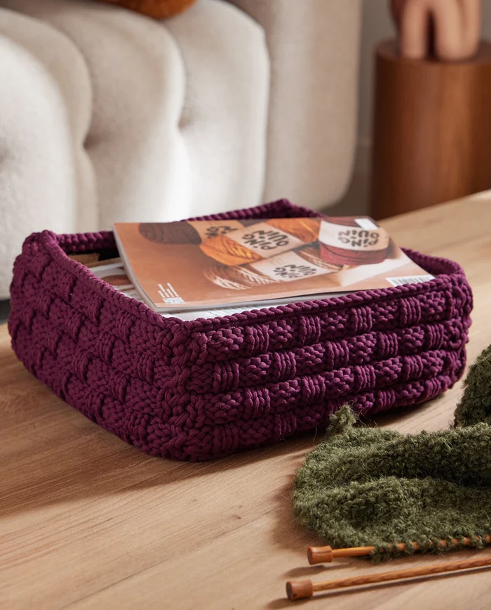 Cesto quadrado roxo trançado com revistas e tecido tricotado verde sobre mesa de madeira