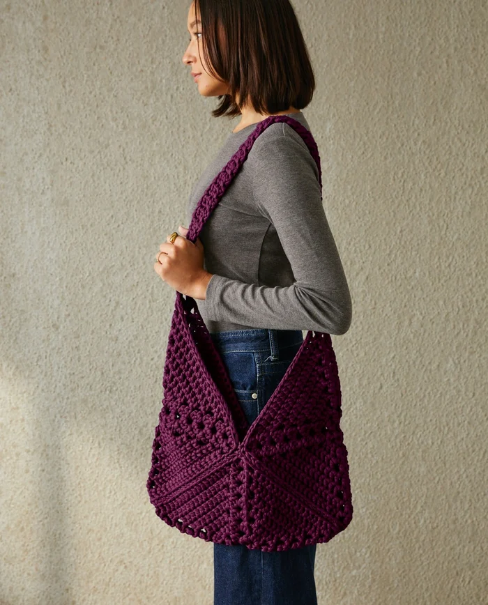 Mulher com mala roxa tricotada ao ombro, parede clara texturada em fundo