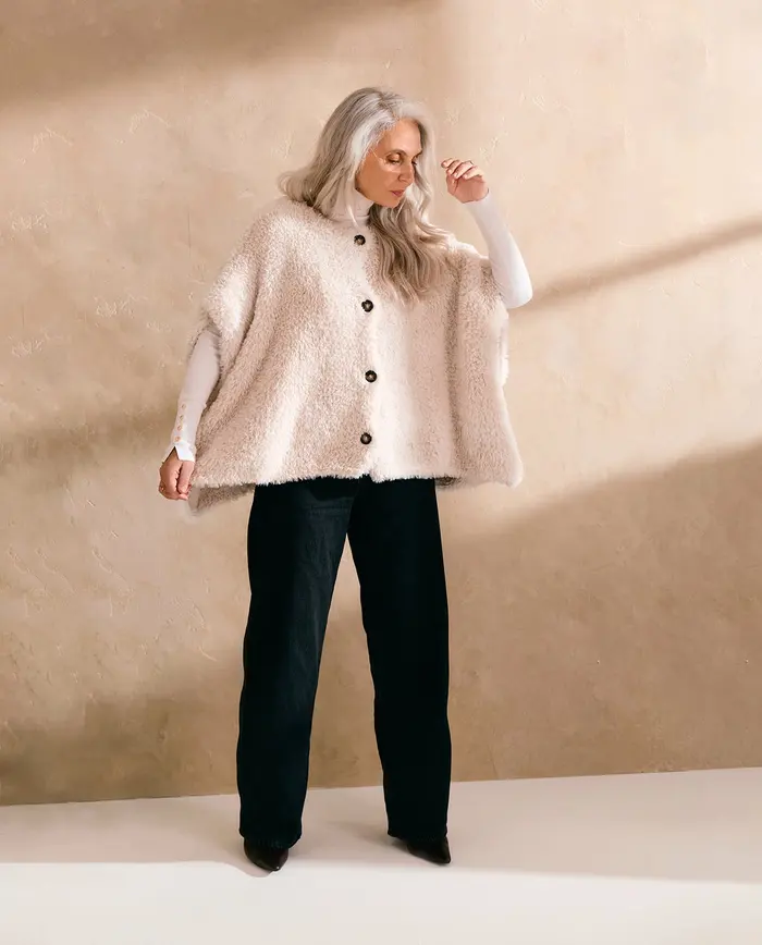 Mulher com capa creme felpuda, camisola branca de gola alta e calças pretas largas