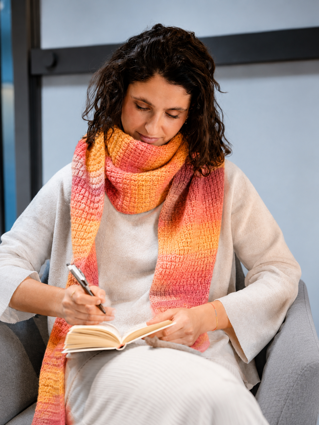 Mulher com cachecol tricotado em cores quentes, sentada a escrever num caderno.