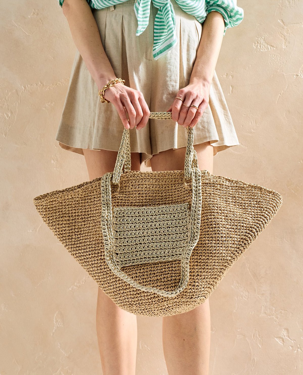 Sacola de ráfia natural com alças e bolso frontal trançados, segurada por mulher com roupas verdes e bege.