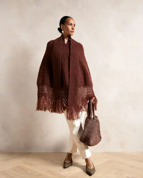 Mulher com capa castanha de franja, calças brancas e mala de malha castanha