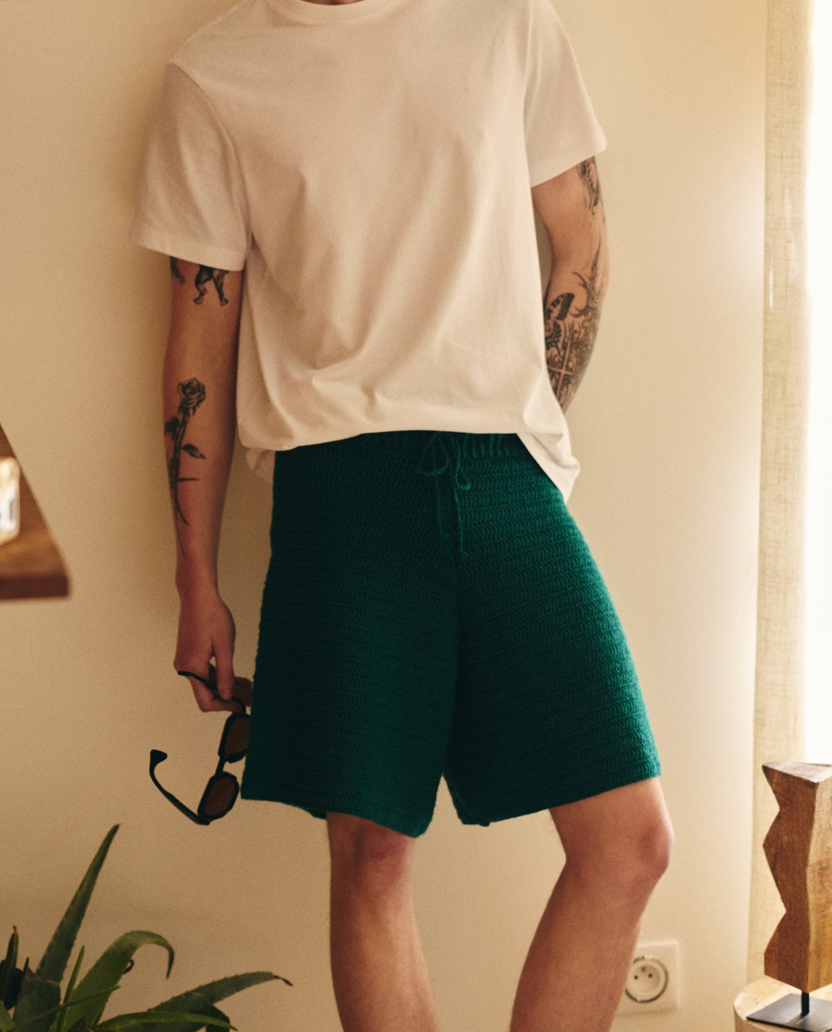 Homem com calções verdes de malha e t-shirt branca, segurando óculos de sol.