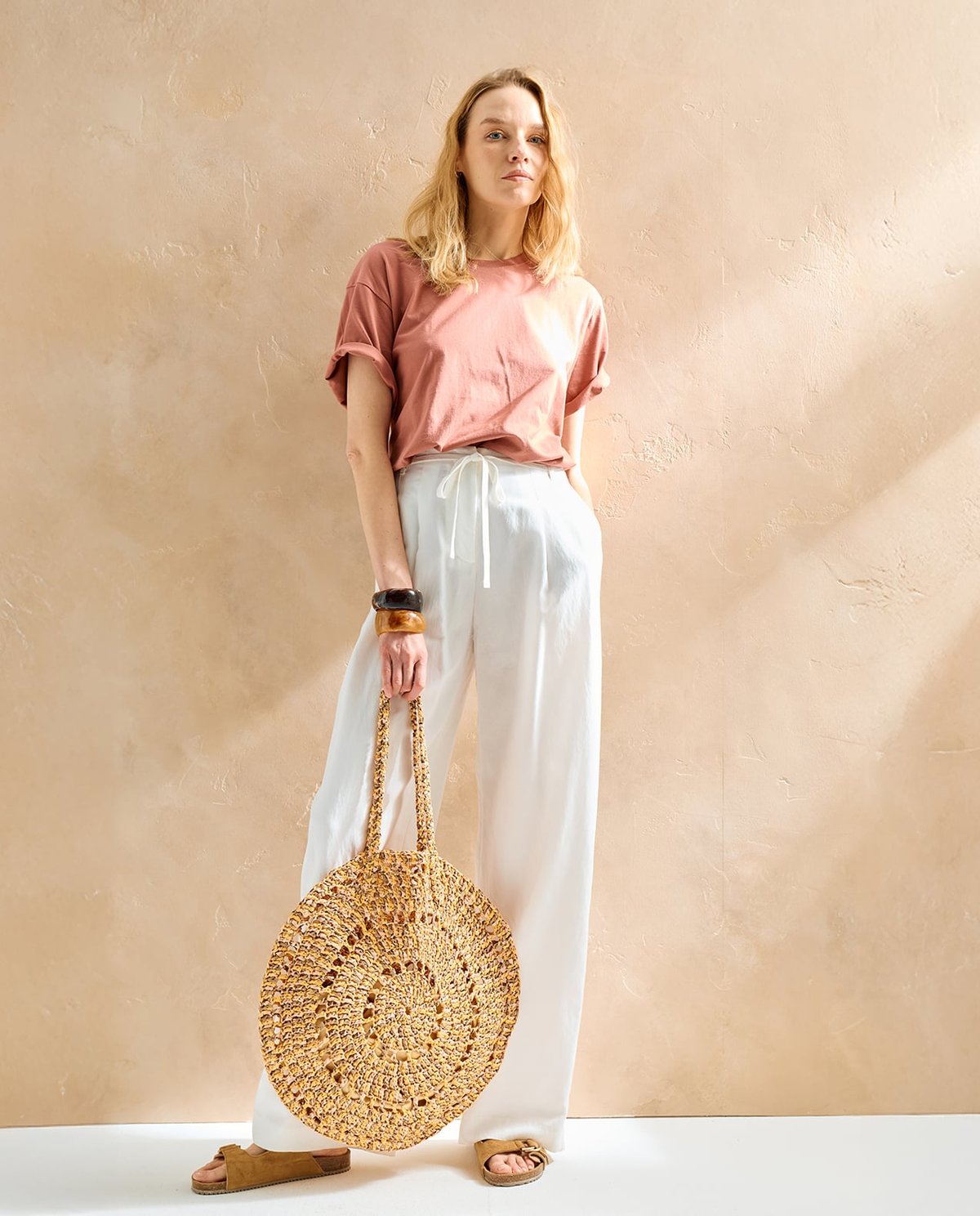 Mulher com camiseta rosa e calças brancas segurando mala redonda de palha