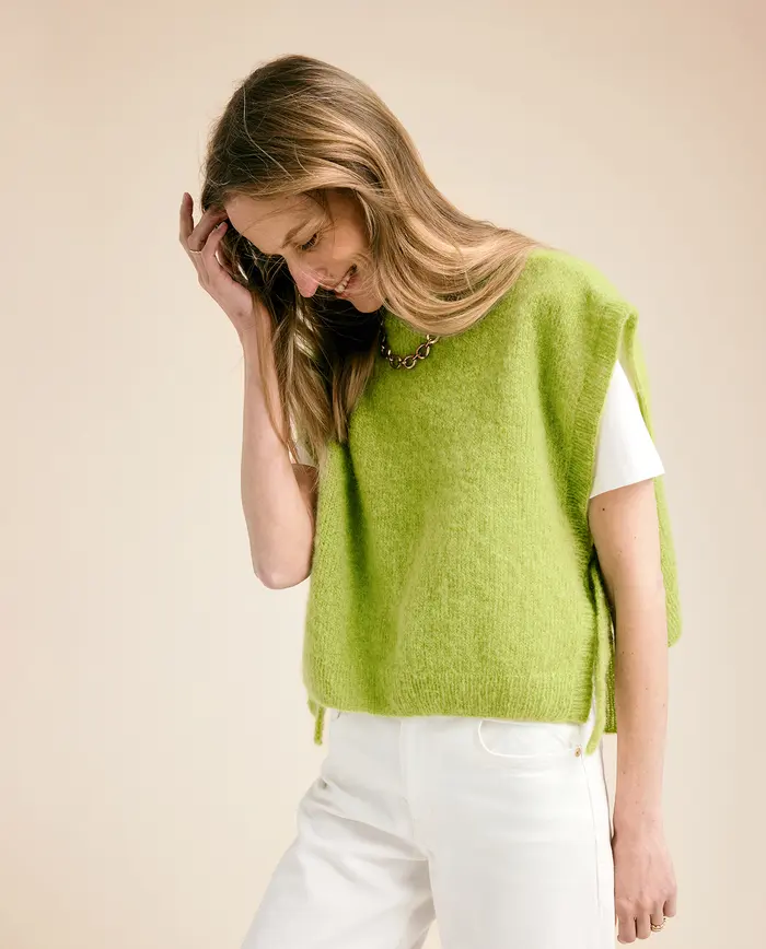 Mulher a vestir top de malha verde claro sobre t-shirt branca e calças brancas, fundo neutro bege.