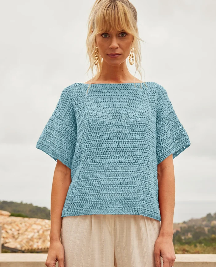 Mulher com camisola de croché azul clara e calças brancas ao ar livre