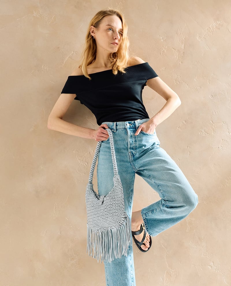 Mulher com top preto, calças de ganga claras, sandálias pretas e bolsa cinzenta de crochet com franjas