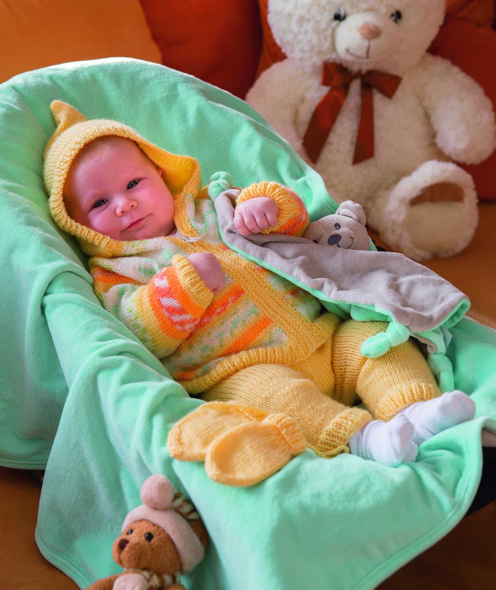 Bebé de malha amarela e laranja deitado numa manta verde com brinquedos de peluche