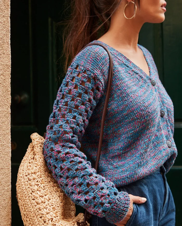 Mulher com cardigan azul-roxo de malha e mochila de palha em fundo de parede e porta escura