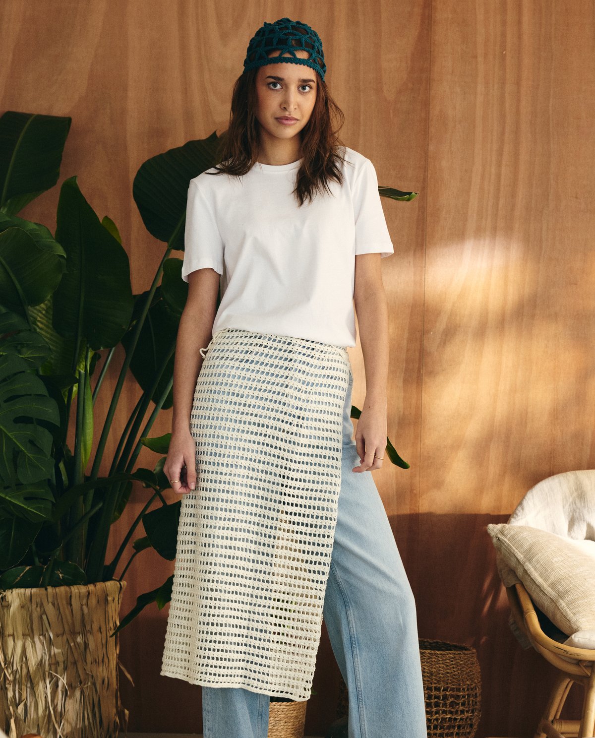 Mulher com camisola branca, saia de malha branca, calças de ganga azul claro e touca verde em ambiente interior com planta e cadeira.