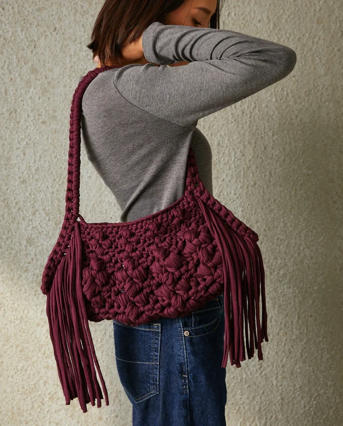 Mala de ombro de croché vinho com franjas largas usada por mulher com camisola cinza e jeans