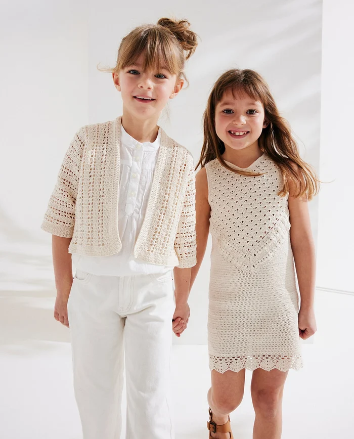 Duas meninas vestindo roupa creme de malha aberta, uma com casaco manga curta e calças brancas, outra com vestido sem mangas, fundo branco.