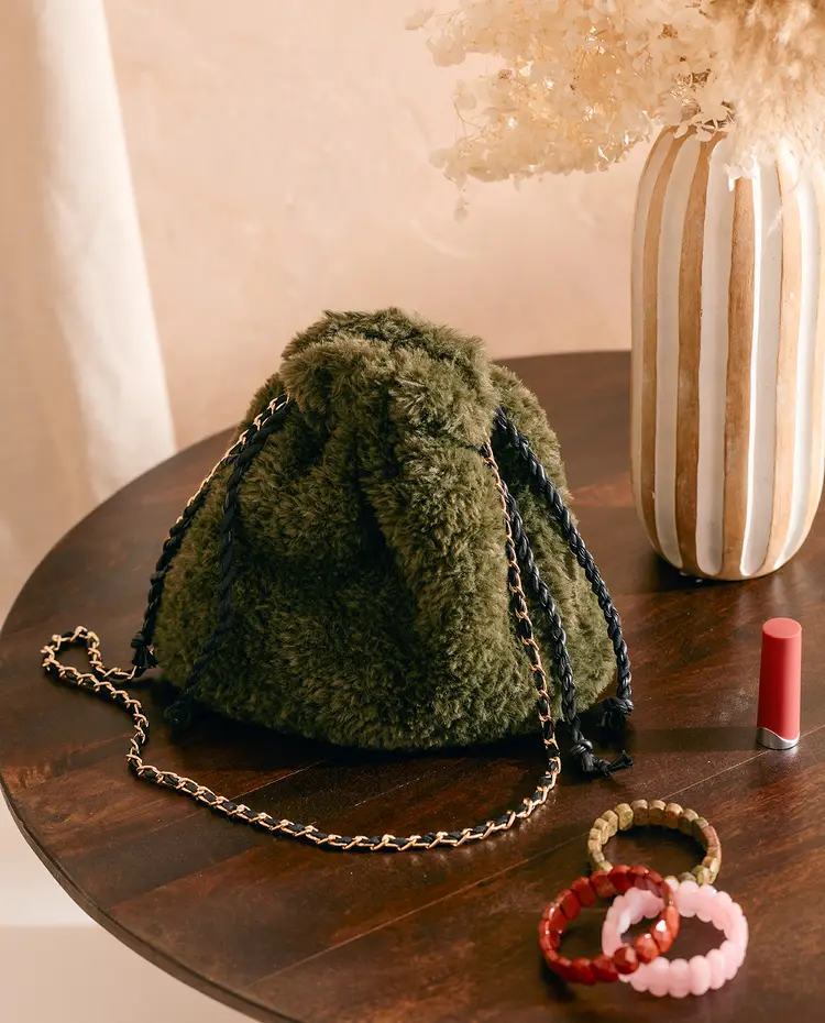 Mala verde felpuda com alça de corrente preta e dourada numa mesa de madeira com batom e pulseiras ao lado
