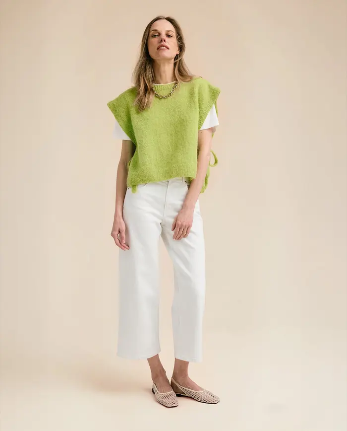 Mulher vestindo colete verde claro, t-shirt branca, calças brancas e sapatos rasos bege em fundo neutro.