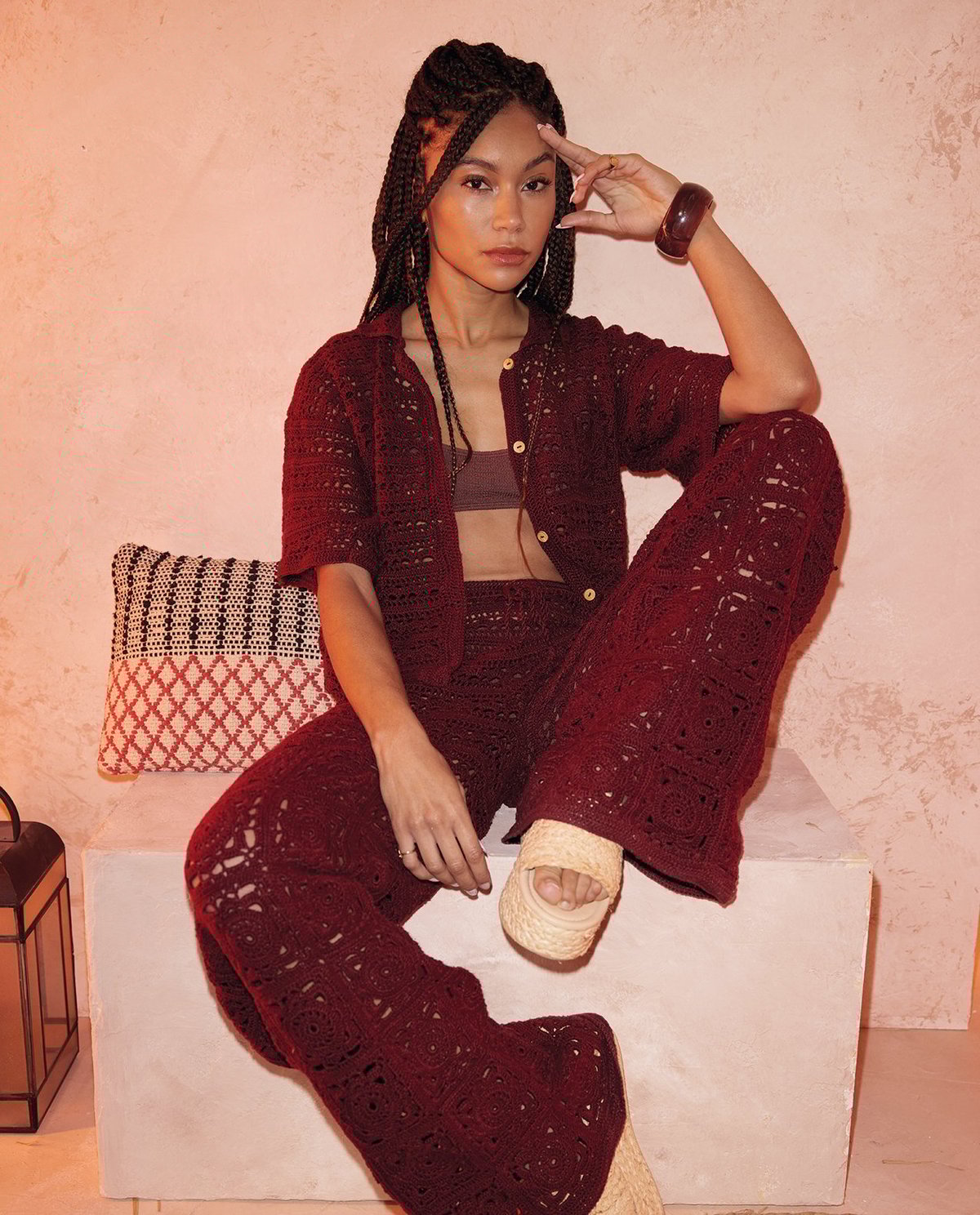 Mulher sentada com conjunto vinho crochetado, top castanho, pulseira castanha e sandálias creme em banco branco com almofada preta, branca e vermelha.