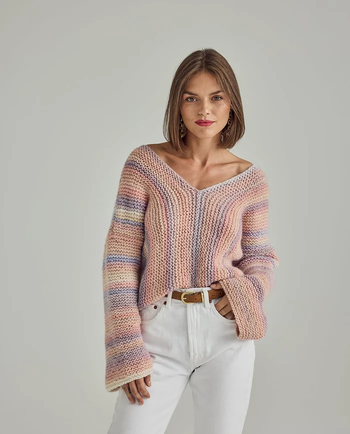 Camisola de malha com padrão riscado em tons pastel, gola em V e mangas largas, vista por mulher com calças brancas e cinto castanho.