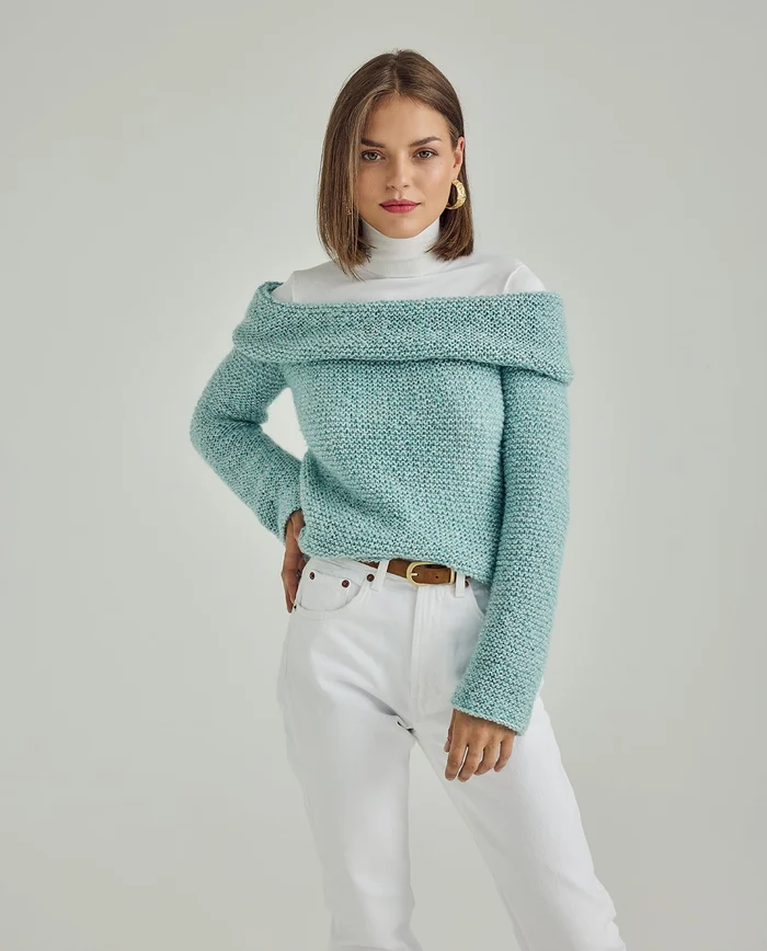 Mulher com camisola azul de malha e calças brancas num fundo cinzento claro.