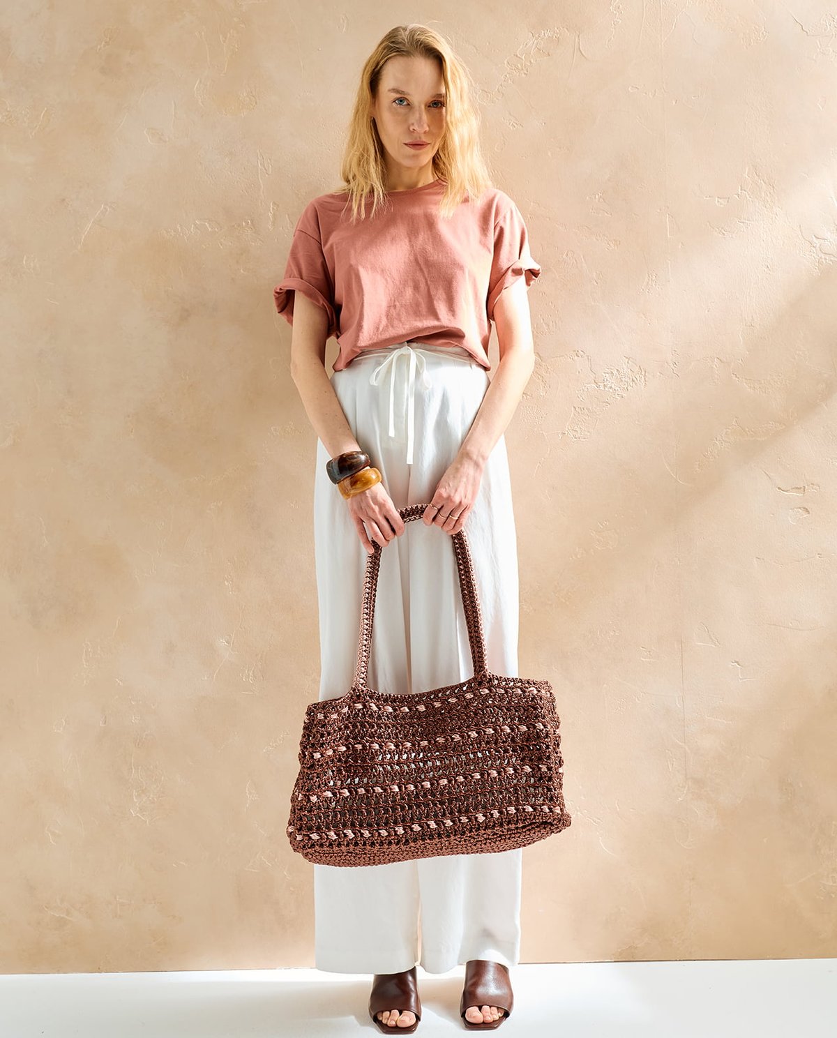 Mulher com mala de crochê castanha, roupa casual em tons rosa e branco, pulseiras e sapatos castanhos