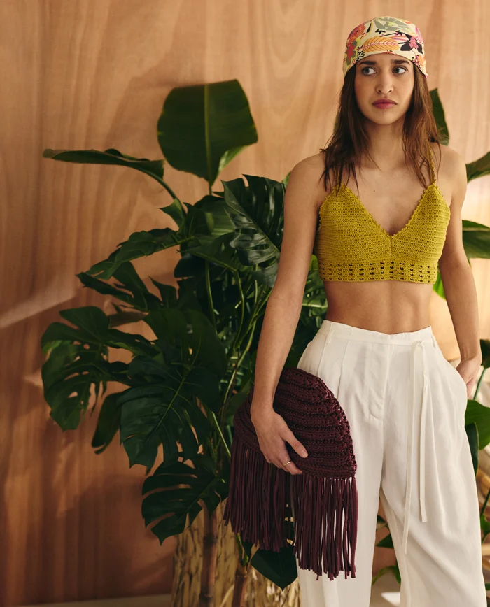 Mulher com top de crochet amarelo, calças brancas e mala de franjas cor de vinho