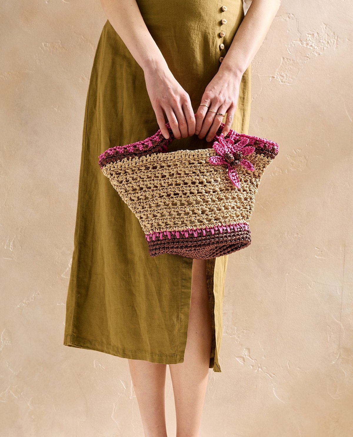Bolsa de palha rosa e castanha com flor, segurada por pessoa com vestido verde-oliva.