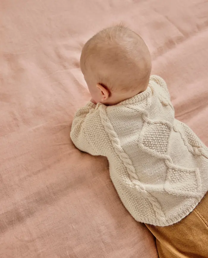 Bebé de costas com camisola de malha branca em superfície rosa.