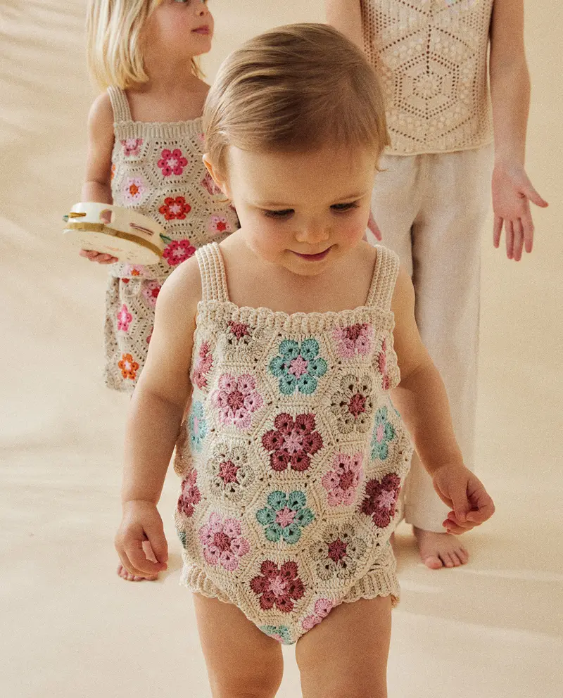 Body de bebé em crochet com flores coloridas, crianças e adulto ao fundo