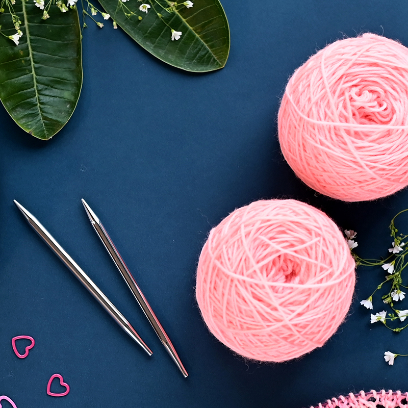 Novelos de lã rosa, agulhas de metal, molas cor-de-rosa em forma de coração, folhas e flores em fundo azul.