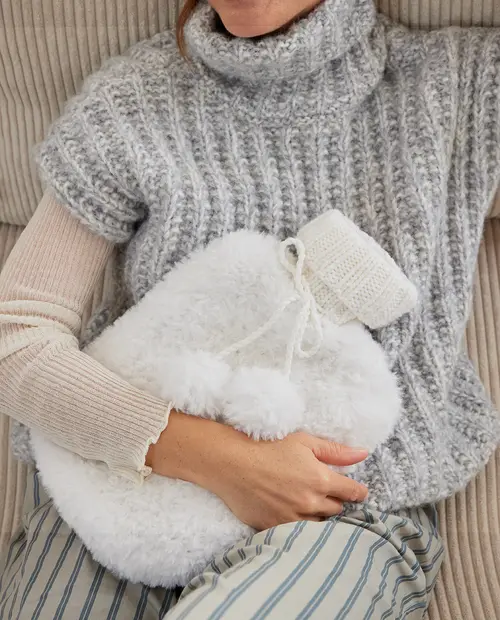 Mala branca felpuda com detalhes em malha branca e pompons, segurada por pessoa vestindo camisola de malha cinzenta e calças às riscas num sofá.