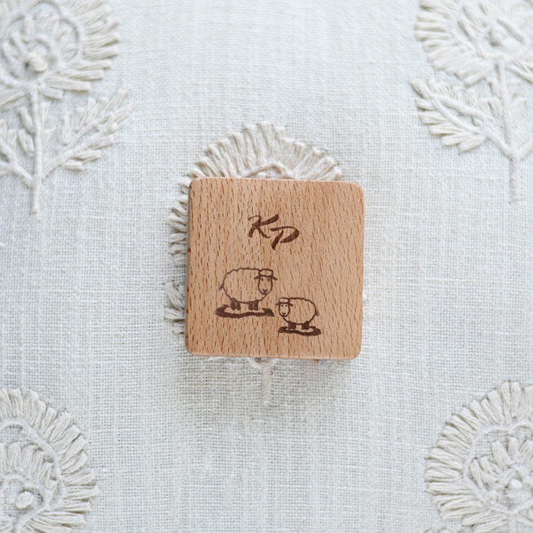Bloco quadrado em madeira clara com ovelhas gravadas e letras KP sobre tecido branco bordado