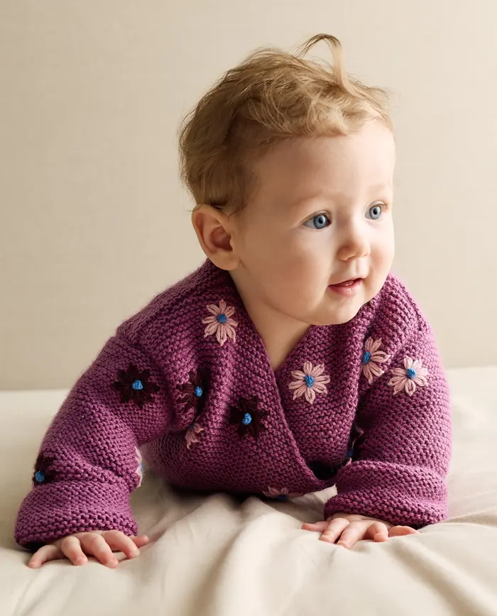 Bebé com casaco roxo bordado com flores numa superfície clara