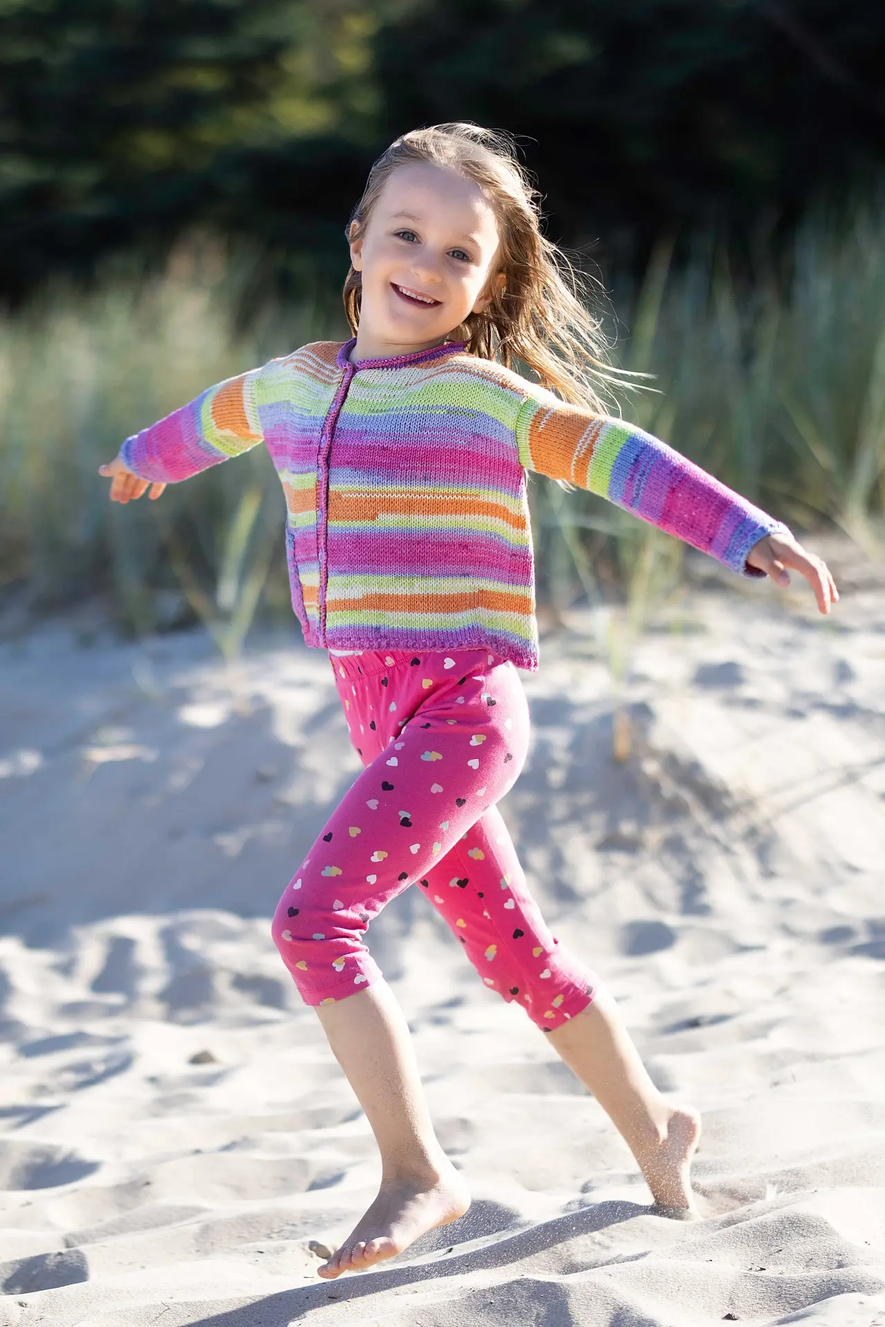 Criança com casaco de malha colorido e leggings rosa estampadas, na areia junto a vegetação