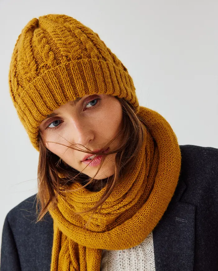 Gorro e cachecol amarelo mostarda de malha com tranças no gorro e cachecol liso, em modelo feminino.