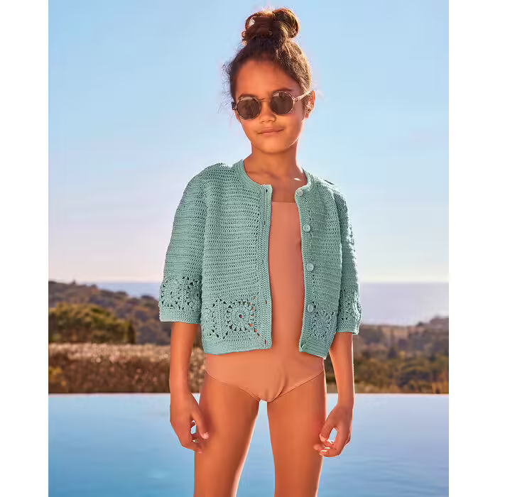 Menina criança com casaco verde-água de malha e óculos de sol junto a piscina