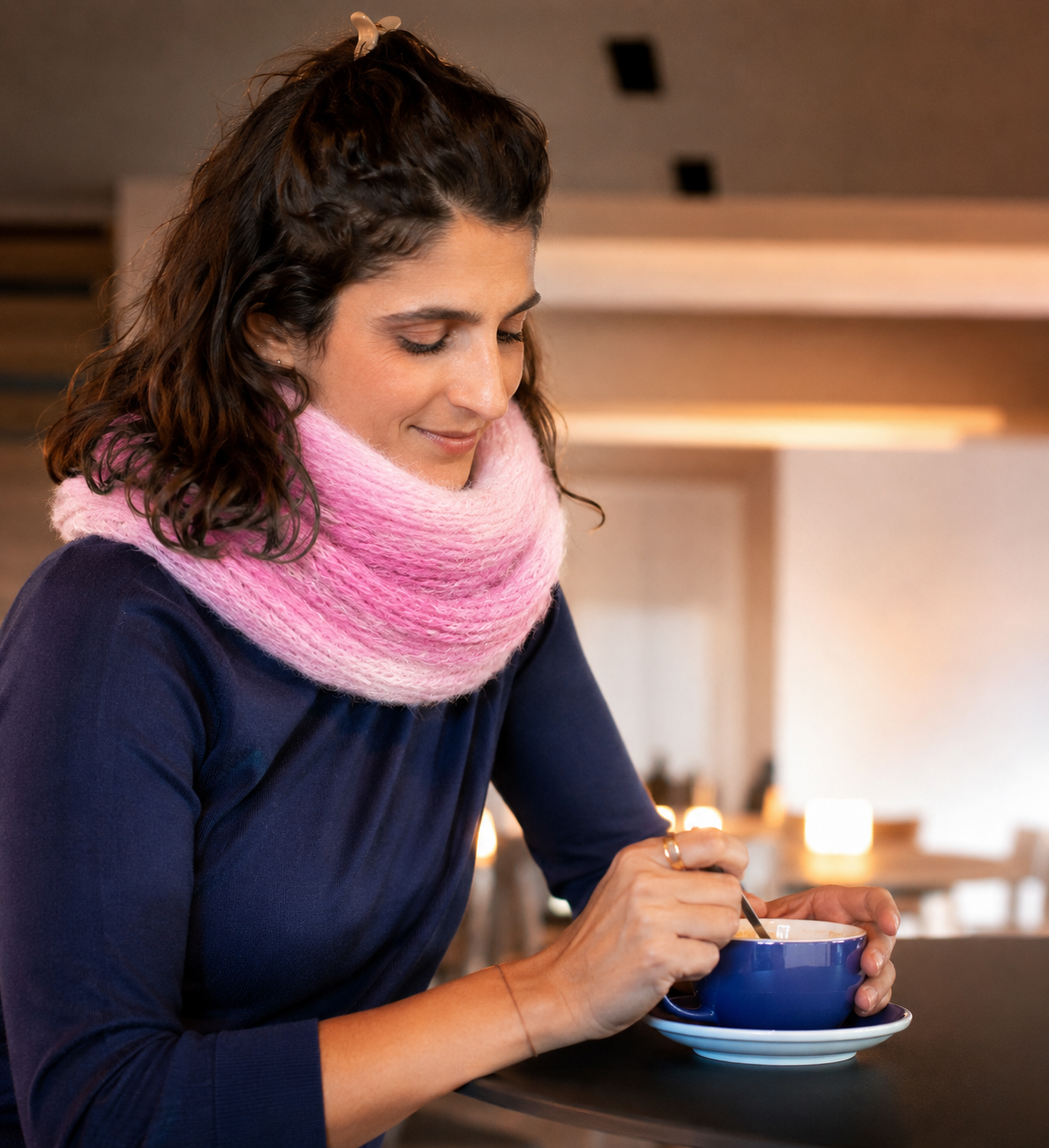Mulher com cachecol rosa e camisa azul segurando chávena azul numa mesa