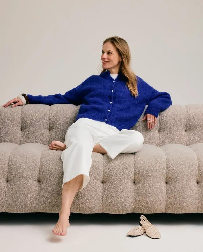 Mulher sentada num sofá cinzento com camisola azul e calças brancas