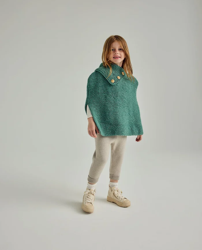 Criança com capa de malha verde, calças creme e botas bege em fundo cinza claro