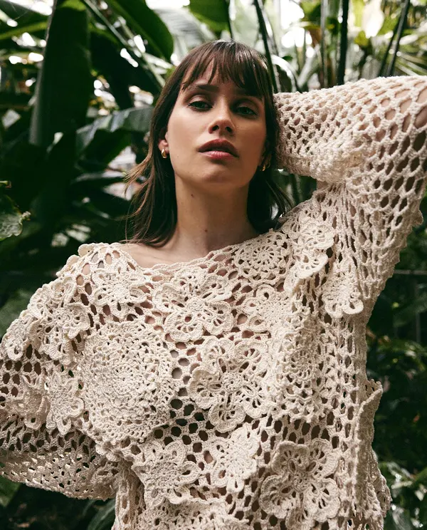 Camisola feminina bege em crochet com padrão floral, usada por modelo junto a plantas tropicais.