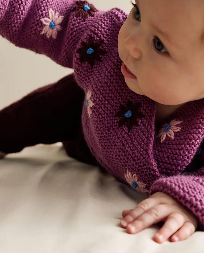 Bebé de pele clara com camisola roxa de malha com flores bordadas
