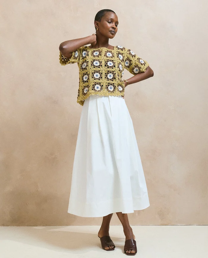Mulher a usar top amarelo de crochet com padrão floral, saia branca comprida e sandálias castanhas em fundo bege.