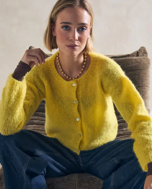 Camisola amarela de malha felpuda com botões brancos e colar dourado, mulher sentada em sofá castanho